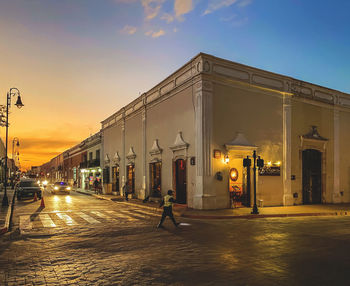 Illuminated street amidst buildings against sky at sunset