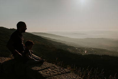 Father and daughter sitting on mountain against sky