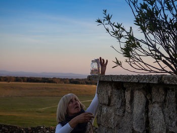 Woman placing jar with illuminated lights on retaining wall against sky during sunset