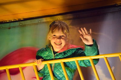 Low angle portrait of cheerful girl waving while standing by railing