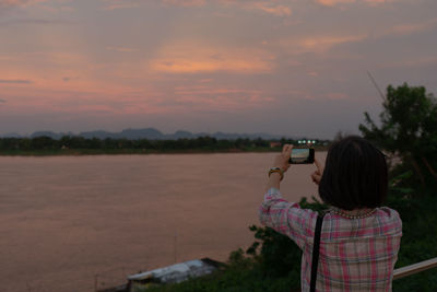 Rear view of woman photographing against sky during sunset