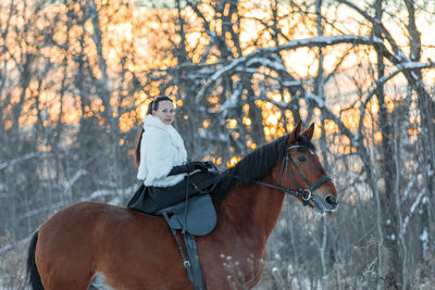 A girl in a white cloak rides a brown horse in winter.