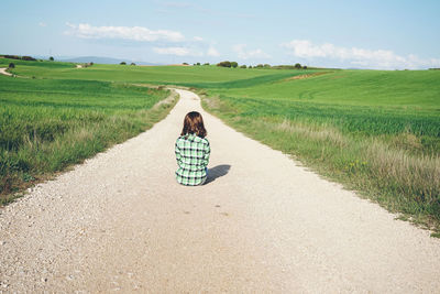 Rear view of woman sitting on dirt road amidst field against sky