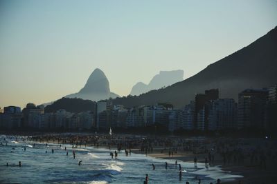 Crowd at beach by city and mountains against clear sky