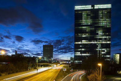 Light trails on city street by buildings against sky at night