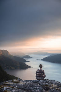 Rear view of woman looking at river amidst mountains against sky during sunset