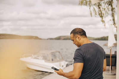 Mature man using smart phone with yacht moored on lake in background