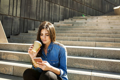 Young woman with coffee to go sitting on stairs looking at cell phone