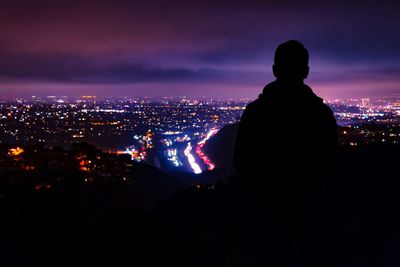 Rear view of silhouette man looking at illuminated cityscape against dramatic sky at night