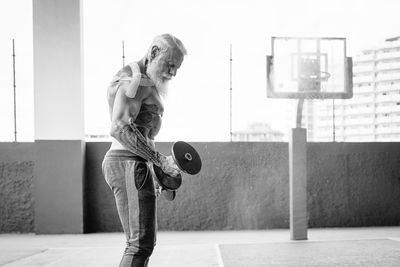 Side view of shirtless man exercising with dumbbell while standing against wall