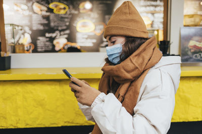 Young woman using smart phone waiting outside cafe in winter during covid-19