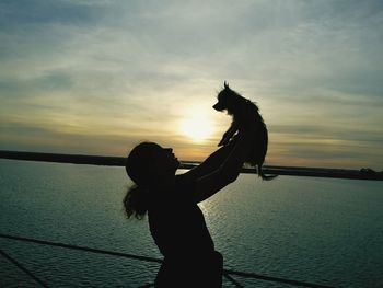 Side view silhouette woman holding dog by sea against sky during sunset