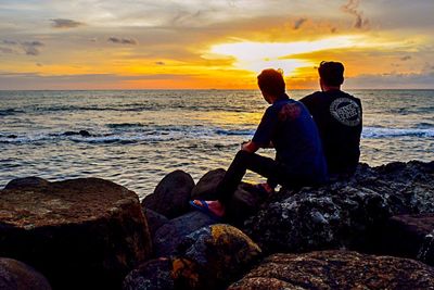 Rear view of men sitting on rock by sea against sky