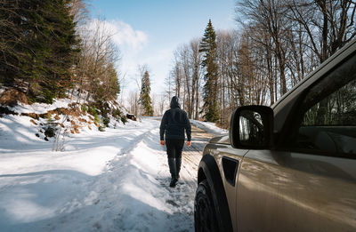 Rear view of man walking on snow covered road by car in forest
