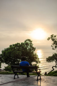 Rear view of man sitting on bench against sky during sunset