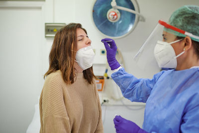 Side view of anonymous medic in protective shield taking analysis from nose of female patient while using medical tool during covid 19 pandemic