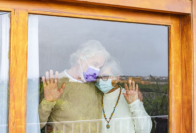 Senior couple wearing mask embracing while standing by window at home