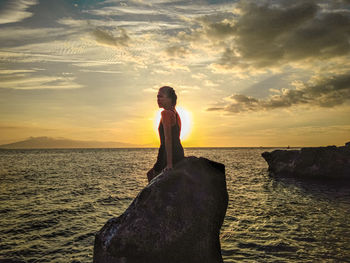 Woman standing on rock by sea against sky during sunset