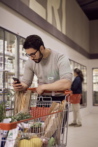 Man text messaging while leaning on shopping cart at refrigerated section in supermarket