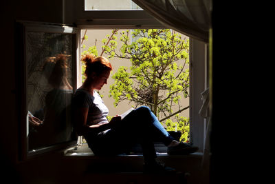 Side view of mid adult woman reading book while sitting on window sill at home