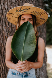 Portrait of woman holding hat while standing against tree