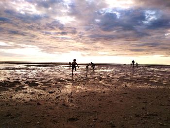 People on shore at beach against cloudy sky during sunset
