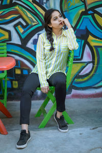 Full length of young woman sitting against graffiti wall