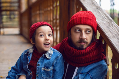 Father and son in red hats and knitted scarves sitting on porch stairs. wooden house logs in autumn