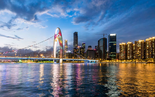 Illuminated bridge over river by buildings against sky