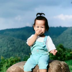 Portrait of cute baby girl sitting on rock against mountain