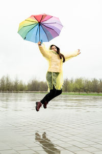 Beautiful brunette woman in yellow raincoat holding rainbow umbrella jumping out in the rain