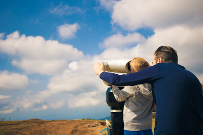 Father and son looking through coin-operated binoculars against sky