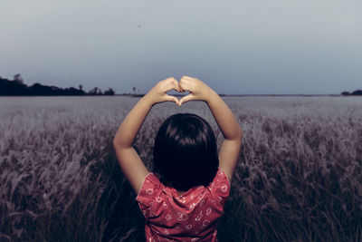Rear view of girl making heart shape while standing on land against clear sky