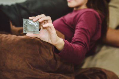 Midsection of woman holding condom packet while lying on bed