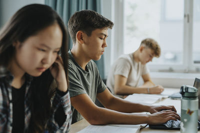 Teenage boy typing while using laptop by girl sitting at desk in classroom