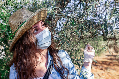 Curious female wearing straw hat and protective mask and gloves touching tree branches while working in farm