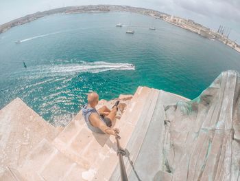 High angle view of man holding monopod while sitting on rock formation against sea