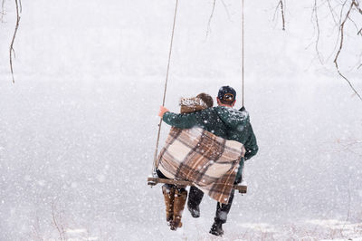 Couple cuddled in blanket look out over river on swing while snowing