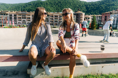 Two young caucasian tanned women are sitting on a bench in a skatepark talking.