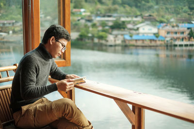Man reading book while sitting on chair against lake