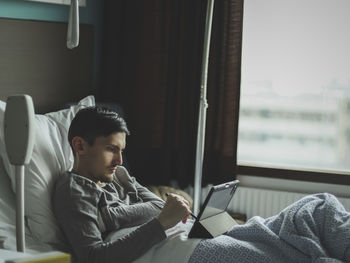 A young man with a tablet lies in bed in a hospital.