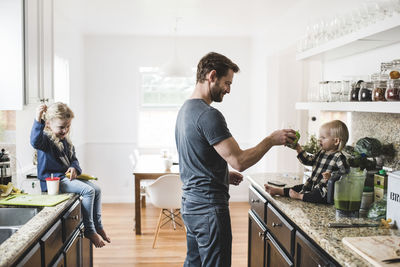 Girl looking at father feeding sister in kitchen at home