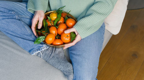 Woman in a warm sweater and jeans holds a bowl of fresh tangerines while sitting on a sofa.