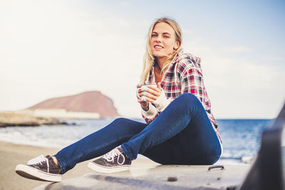 Portrait of smiling young woman holding mug while sitting on off-road vehicle against sea