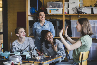 Smiling female student gesturing while sitting with friends and teacher in classroom at high school