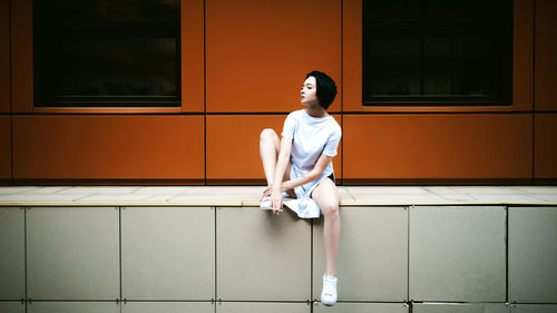 Full length of young woman sitting on walkway against orange building