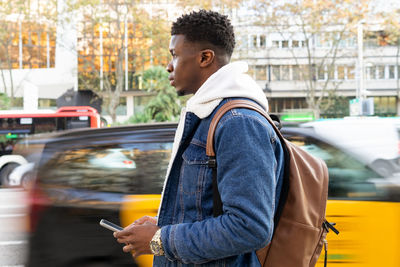 Side view of pensive african american male with curly hair in trendy outfit walking with smartphone in hand against driving car on city street in daytime