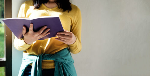 Midsection of woman reading book while standing against wall