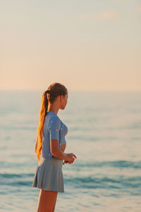 Young woman standing at beach against sky during sunset