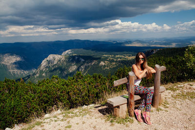 Full length portrait of woman sitting on bench against landscape